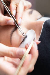 Crop professional female master with tweezer applying artificial eyelashes on young female client in face mask in light modern salon - ADSF22698