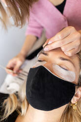 Crop professionelle weibliche Meister mit Pinzette Anwendung künstliche Wimpern auf junge weibliche Kunden in Gesichtsmaske in hellen modernen Salon - ADSF22700