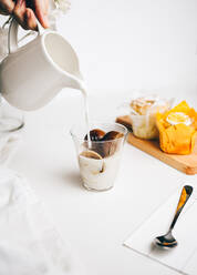 Crop anonyme Person, die heiße Milch in ein Glas mit köstlichen schmelzenden Schokoladenbomben auf dem Tisch neben süßen Cupcakes - ADSF22701