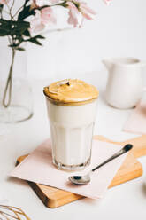 Teelöffel mit süßem Schaum über frischem, leckerem Milchkaffee, serviert auf einem hölzernen Schneidebrett in einer modernen, hellen Küche - ADSF22705