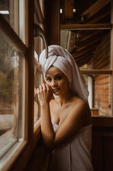 Young gentle female in towels touching face while looking at camera in wooden cabin - ADSF22713