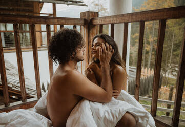 Romantic young undressed couple cuddling gently while sitting on wooden cottage porch and covering bodies with cozy white blanket on autumn weather - ADSF22721