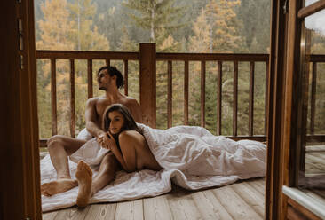 Romantic undressed couple lying on cozy blanket and cuddling on wooden cottage terrace against deciduous forest on autumn - ADSF22722