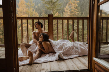 Romantic undressed couple lying on cozy blanket and cuddling on wooden cottage terrace against deciduous forest on autumn - ADSF22723