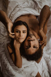 Von oben romantisches junges Paar, das Rücken an Rücken sitzt und die Köpfe auf den Schultern des anderen ruhen lässt, während es auf einer weichen Decke entspannt in die Kamera schaut - ADSF22728
