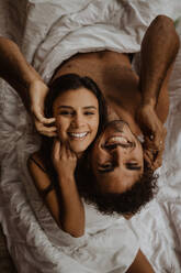 Von oben fröhliches, unbekleidetes Paar, das Rücken an Rücken auf einer gemütlichen Decke sitzt und mit einem breiten Grinsen in die Kamera schaut - ADSF22729