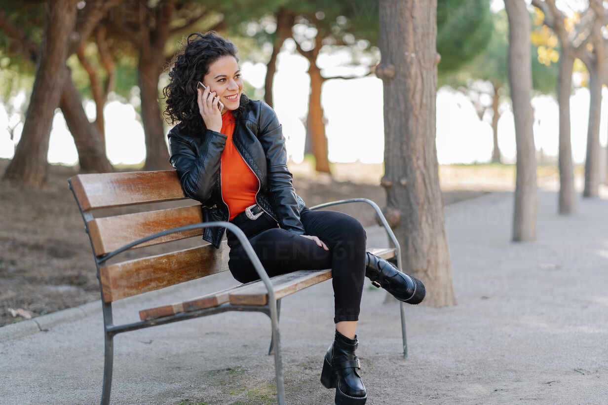 Happy pretty female in casual clothes and leather jacket having  conversation via mobile phone and looking away with toothy smile while  sitting on a bench in the street stock photo