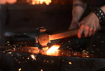 Seitenansicht eines jungen, brutalen, bärtigen Handwerkers in Schürze und Schutzbrille, der während der Arbeit in einer traditionellen Schmiede Eisen auf einem Amboss mit einem Hammer erhitzt und schlägt - ADSF22749