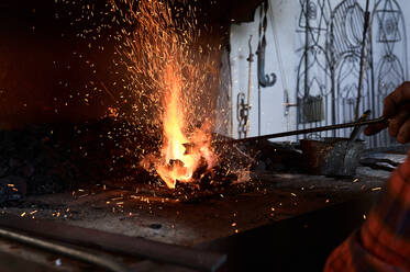 Unkenntlich gemachter männlicher Schmied, der einen Metallstab auf einer brennenden Flamme erhitzt, während eines Schmiedevorgangs in einer Werkstatt - ADSF22754