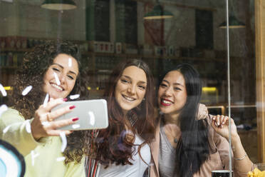 Friends taking selfie on mobile phone by cafe window - PNAF01397