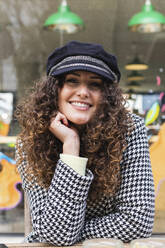 Lächelnde Frau mit lockigem Haar und Mütze sitzt mit Hand am Kinn in einem Straßencafé - PNAF01413