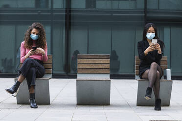 Young women wearing protective face mask using mobile phone while sitting with social distancing on bench - PNAF01423