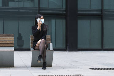 Woman with face mask talking on mobile phone while sitting on bench - PNAF01424