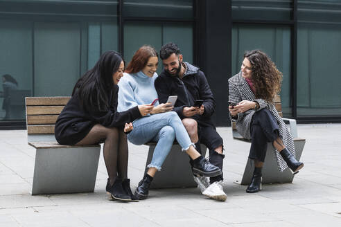 Junger Mann und Frauen benutzen ein Mobiltelefon, während sie auf einer Bank sitzen - PNAF01427