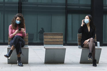 Woman wearing face mask using mobile phone while sitting on social distance with female over footpath - PNAF01453