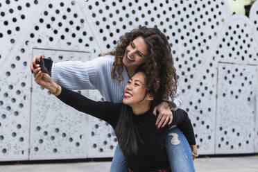 Happy young female friends taking selfie through smart phone in front of wall - PNAF01460