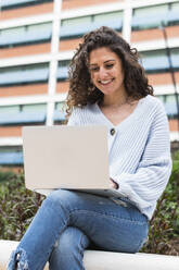 Smiling woman using laptop while sitting on retaining wall - PNAF01467