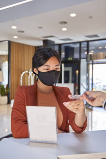 Frau mit Gesichtsmaske, die an der Hotelrezeption den Kartenschlüssel von der Rezeptionistin entgegennimmt - VEGF04208