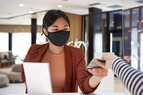 Junge Frau mit Gesichtsmaske, die den Kartenschlüssel von der Rezeptionistin im Hotel entgegennimmt - VEGF04209