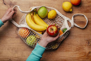 Wiederverwendbare Baumwoll-Netztasche mit frischen Früchten und der Hand eines Mannes, der einen reifen Apfel hält - RTBF01595