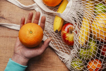 Reusable cotton mesh bag with fresh fruits and hand of man holding ripe orange - RTBF01597