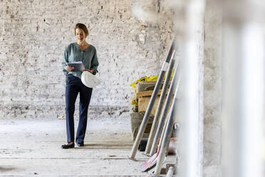 Architektin mit Schutzhelm schaut auf ein digitales Tablet, während sie auf einer Baustelle steht - PESF02758