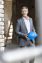 Männlicher Architekt, der einen Schutzhelm hält, während er auf einer Baustelle steht - PESF02770