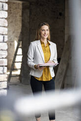 Smiling female architect holding notebooks at construction site - PESF02773