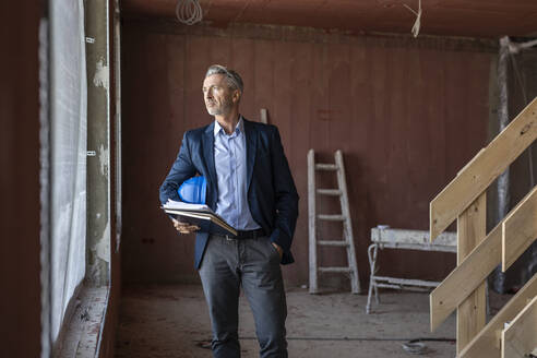 Männlicher Architekt mit Hand in der Tasche, der ein Notizbuch hält und durch ein Fenster auf eine Baustelle blickt - PESF02787