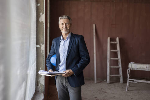 Lächelnder männlicher Architekt, der einen Schutzhelm hält, während er in der Nähe eines Fensters auf einer Baustelle steht - PESF02788