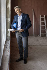 Male architect looking at documents while standing near window at site - PESF02790