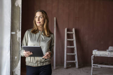 Female architect looking away while holding digital tablet near window at site - PESF02795