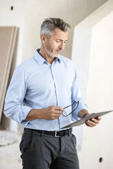 Male architect holding eyeglasses while looking at digital tablet at construction site - PESF02803