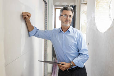 Male professional with digital tablet leaning on wall in corridor at construction site - PESF02814