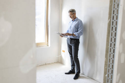 Männlicher Unternehmer, der ein digitales Tablet benutzt, während er an einer Wand auf einer Baustelle steht - PESF02815