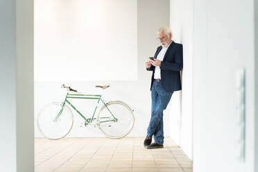 Ein älterer männlicher Unternehmer benutzt ein Smartphone, während er sich im Büro an die Wand lehnt - MOEF03692
