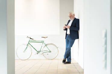 Senior male entrepreneur using smart phone while leaning on wall at office - MOEF03692