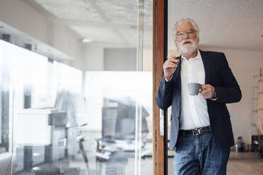 Älterer Geschäftsmann hält Zigarette mit Kaffeetasse, während er sich an eine Glaswand im Büro lehnt - MOEF03693
