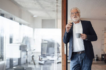 Senior businessman holding cigarette with coffee cup while leaning on glass wall at office - MOEF03693