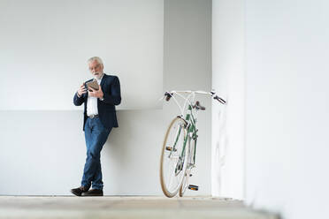 Businessman using digital tablet near bicycle while leaning on wall at office - MOEF03696