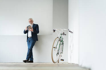 Geschäftsmann, der ein digitales Tablet in der Nähe eines Fahrrads benutzt, während er sich im Büro an die Wand lehnt - MOEF03696