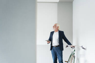 Älterer männlicher Fachmann, der wegschaut, während er in der Nähe eines Fahrrads im Büro steht - MOEF03697