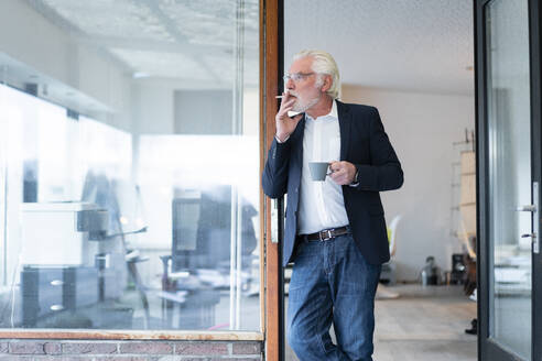 Älterer Geschäftsmann raucht Zigarette und lehnt sich an eine Glaswand im Büro - MOEF03714
