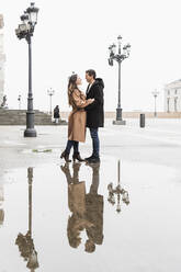 Glücklicher, fröhlicher Mann steht und umarmt seine hübsche Freundin in einem warmen Mantel, während sie zusammen auf der Straße an einem klaren Herbsttag stehen - ADSF22784