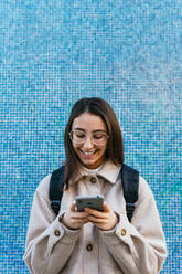 Crop attractive smiling female student in warm wear with backpack using modern mobile phone on sidewalk on street - ADSF22788