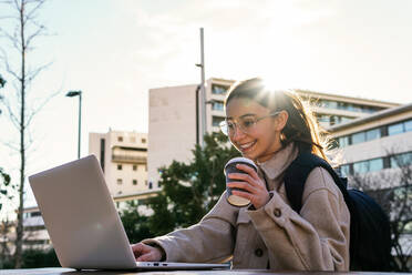 Glücklich lächelnde Studentin in warmem Mantel und Brille mit Rucksack, die an einer Hausarbeit auf einem Netbook arbeitet, während sie an einem Tisch mit Kaffee zum Mitnehmen in einem sonnigen Park sitzt - ADSF22792