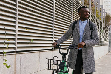 Young content African American male employee in coat with bike standing on urban pavement against ribbed wall and looking away - ADSF22799