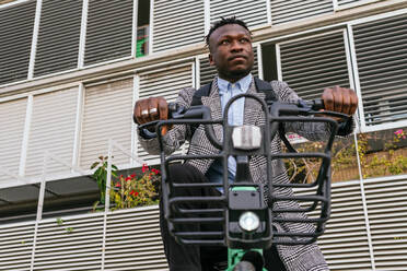 Junger zufriedener afroamerikanischer männlicher Angestellter im Mantel mit Fahrrad, der auf einem städtischen Bürgersteig vor einer gerippten Wand steht - ADSF22801