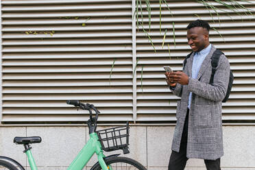 Niedriger Winkel des Inhalts afroamerikanischer männlicher Manager, der in der Nähe eines Fahrrads in der Stadt Textnachrichten auf einem Handy schreibt - ADSF22802