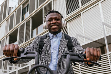 Young content African American male employee in coat with bike standing on urban pavement against ribbed wall - ADSF22804